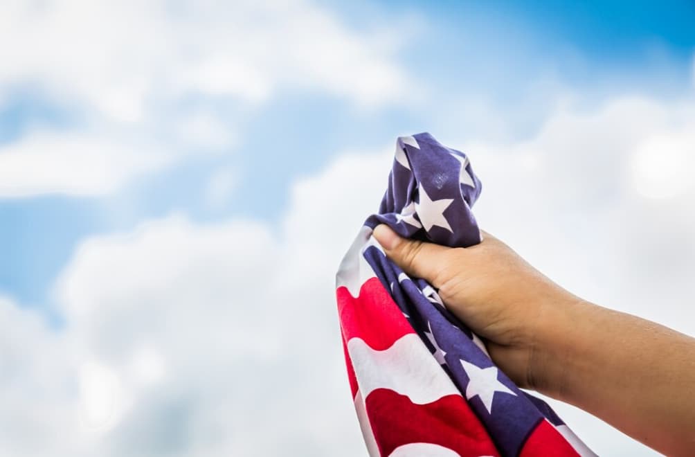 A hand holding the American flag against a cloudy sky