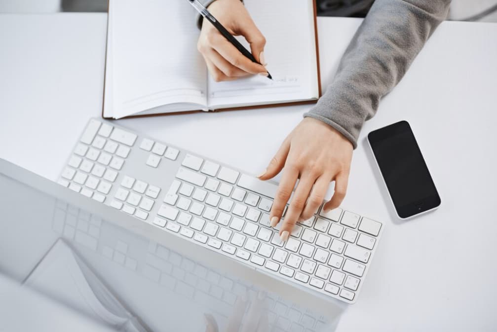 A person multitasks with a keyboard, notepad, and smartphone